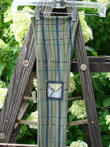 Image alt text: Barclay Patched Scarf with linear green olive plaid pattern and rolled-up edges on a wooden ladder