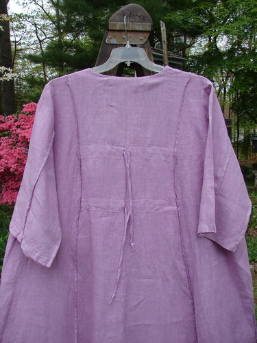 Barclay Linen Double Tie Back Jacket Garden Side Lilac Size 2 on a wooden swing, showcasing deep V neckline, A-line sweep, vertical stitching, drop pockets, varying hemline, and garden-themed paint.