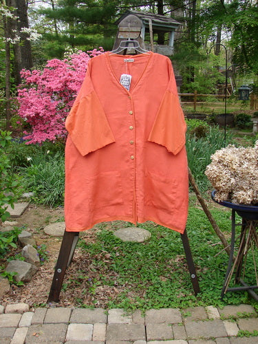 Barclay NWT Linen Cotton Sleeve Pocket Cardigan in Clementine displayed on a rack, featuring wooden-like buttons, upward curved hemline, drop shoulders, and squared-off pockets.