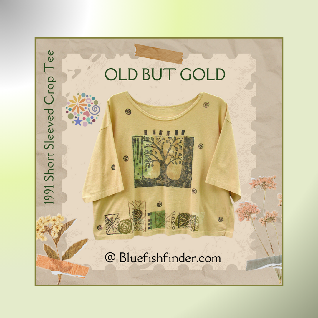 A unique graphic design on a yellow cropped tee shirt from 1991 text stating, "Old But Gold.", A Blend of tans support the background in a pastel gradient matting The Blue Fish Finder's Circle logo and text surround the edges with dried flower accents.