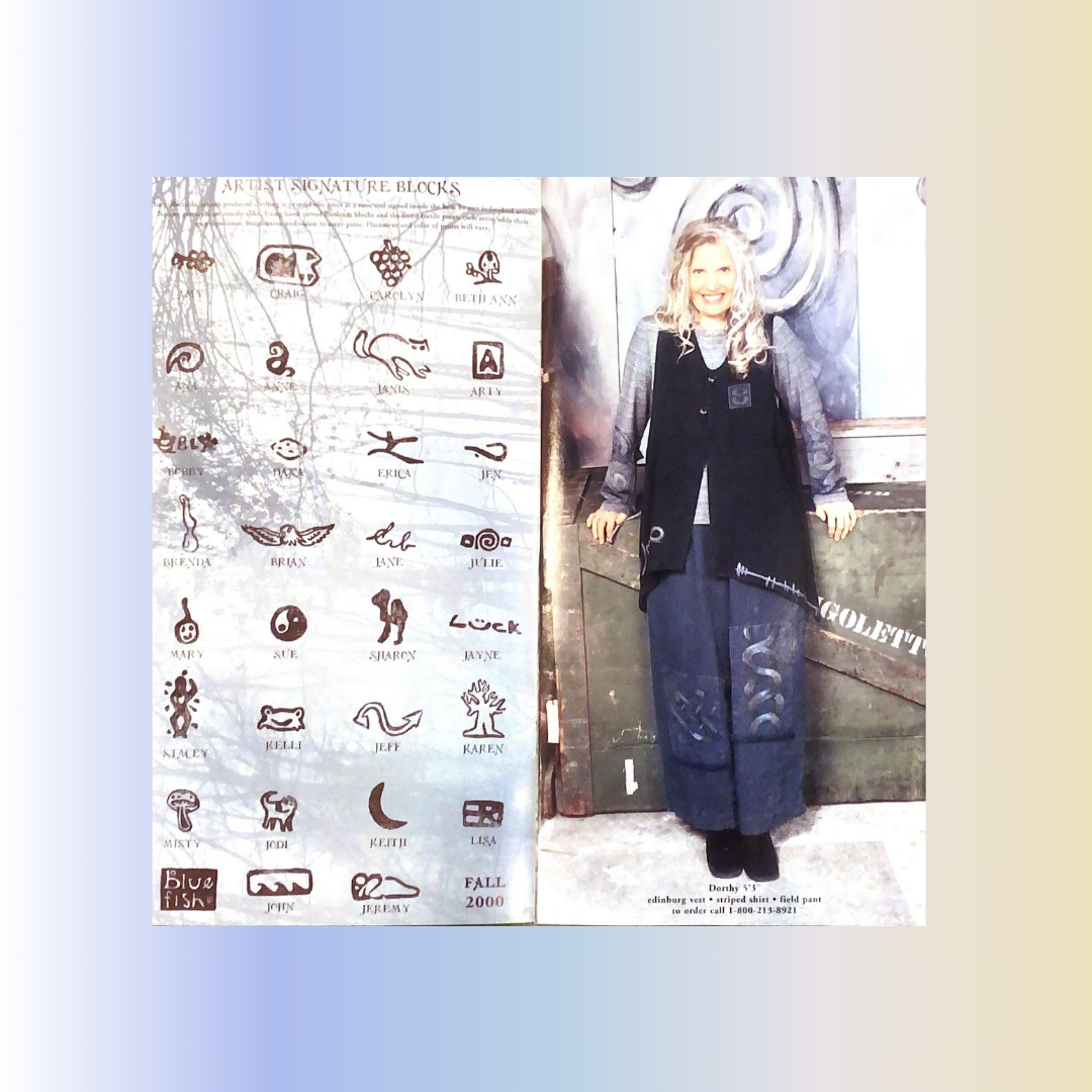 A woman standing next to a poster of artist signatures, wearing a Black Edinburgh vest and mated hemp field pant, surrounded by the catalog cover for the 2000 year and Fall season. Rediscover vintage Blue Fish clothing at Bluefishfinder.com.