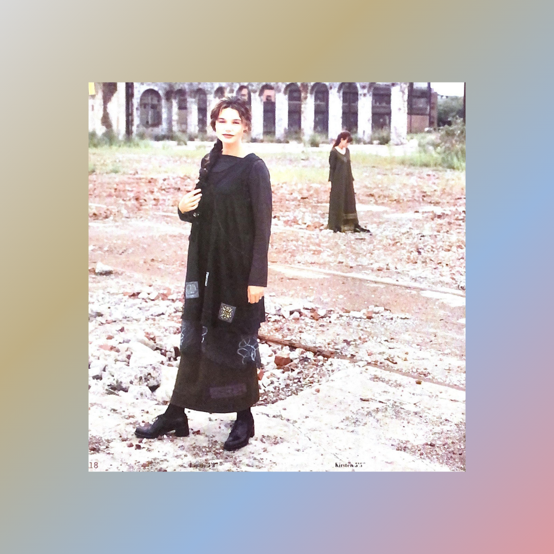 A calm image of a model displaying the Rayon Lycra Rib collection for 2000 Holiday. She wears a jumper top a skirt and a mid layer of silk organza. Her braid is to the side and she stands on a dirt road with a majestic building and a far away friend.