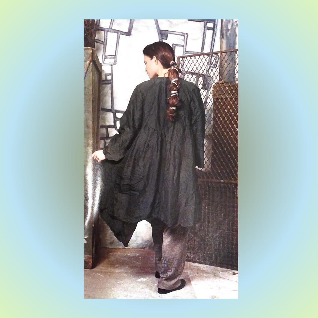 A Model in a studio shot, from the transitions 2000 collection poses with her back to the camera sporting the Shaunting silk Wrap jacket and drawstring pant collection from 2000. She has a long single twisted braid with gray ribbon. There is a wire gate.