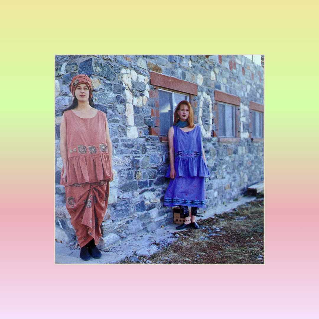 Two models stand against a stone wall in sleeveless dresses called the 1991 Button Tier. Each displaying a peplum and straight skirt appearing as two pieces, but really a single dress with a detachable lower portion. One skirt is gathered by hand.