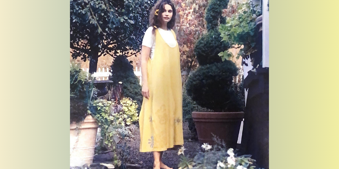 A Model with a long side braid, stands in a garden featuring a large topiary in the background. She is barefooted on a slate step, wears a yellow jumper with paint around the hem and a creme under piece. Large potted plants are in the foreground.