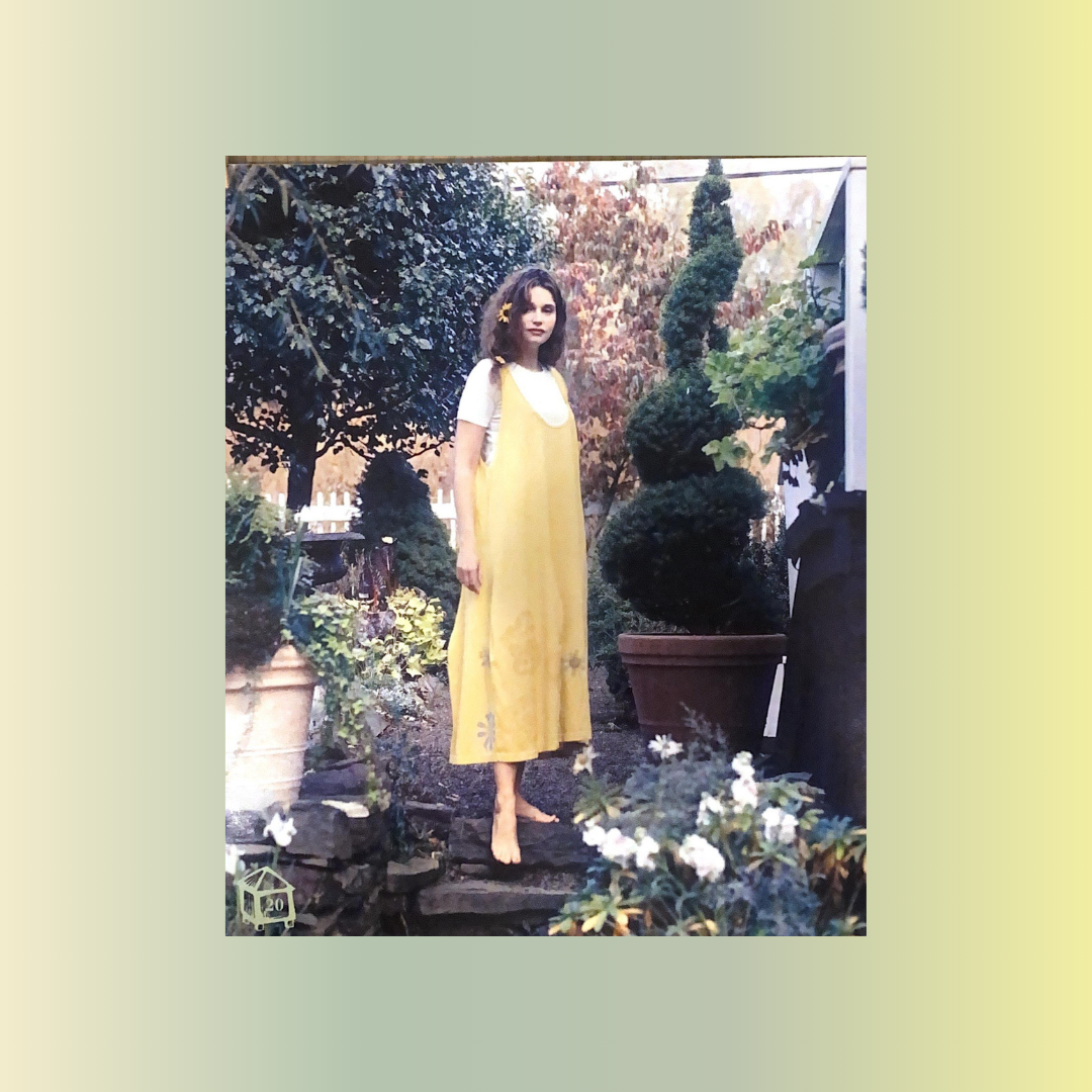 A Model with a long side braid, stands in a garden featuring a large topiary in the background. She is barefooted on a slate step, wears a yellow jumper with paint around the hem and a creme under piece. Large potted plants are in the foreground.