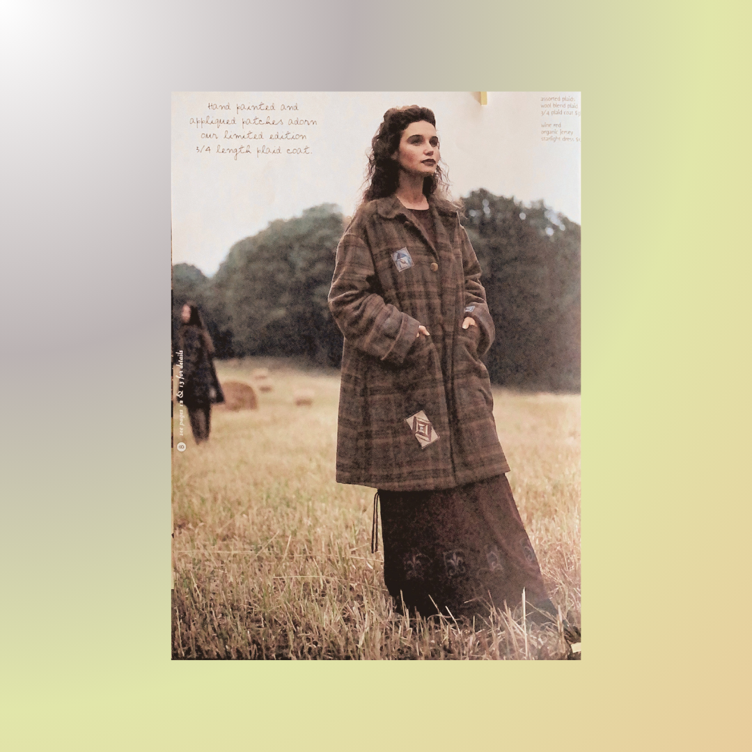 A woman in a long dress and a car length wool plaid coat stands in a field, showcasing a vintage Blue Fish Coat from the 1999 collection.