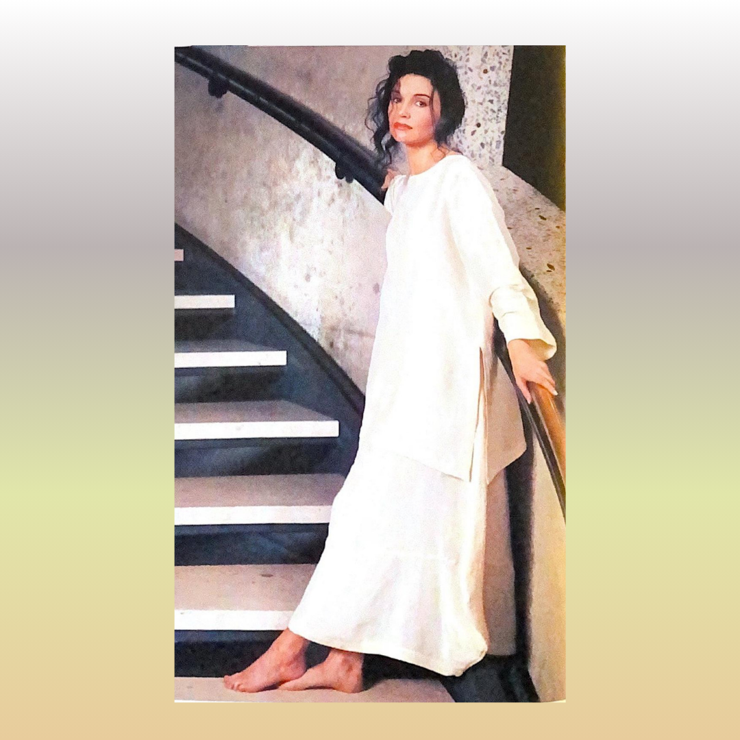 At the bottom of a curved stairway, leaning against the rail, A woman wears a white silk duo from the Summer collection of 1998, a Vintage Blue Fish Clothing outfit for a fancier occasion.