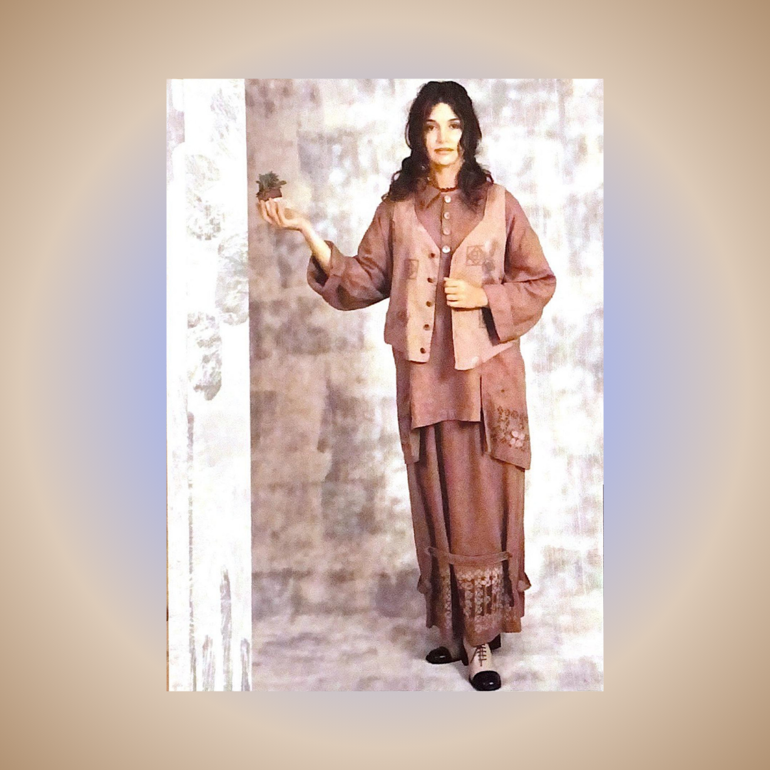 A Model displays a layered look in the color Carmel from the Spring Collection of 2000. She is wearing the Side Tab Skirt, The painters smock and the Hemp crop vest. She holds one hand up in a studio shot with a mottled backdrop in gray and white.