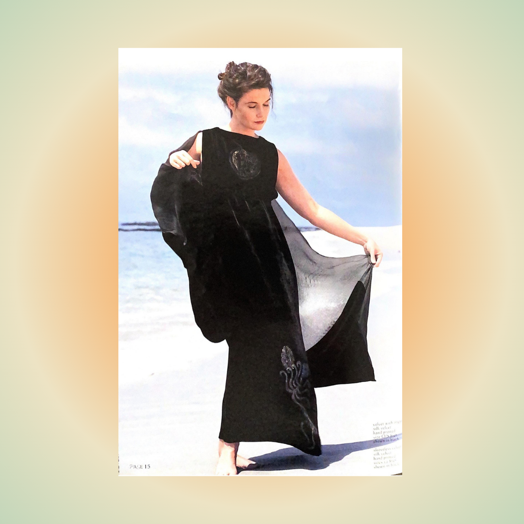 A woman in a black velvet painted pencil dress with her eyes lowered, sweeps a matching velvet off the shoulder shawl on a shoreline with the wind moving her lower skirt.