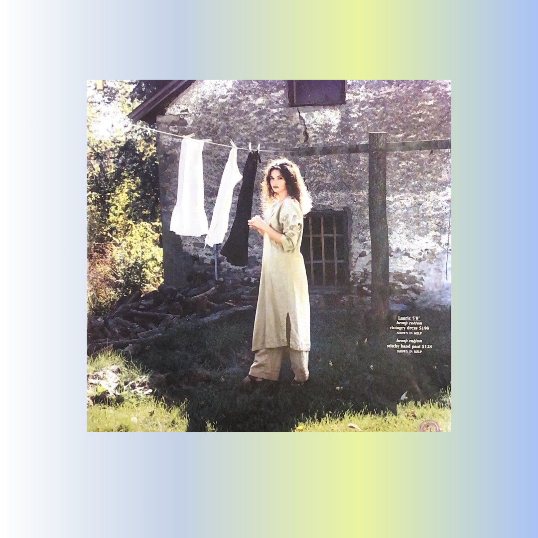 A woman stands in a grassy sunny area hanging clothing on a line. The shadows create a juxtaposition in mood and theme. A stone home is in the background with a wooden framed windows. She is exhibiting 2001 spring clothing for BFC last year.