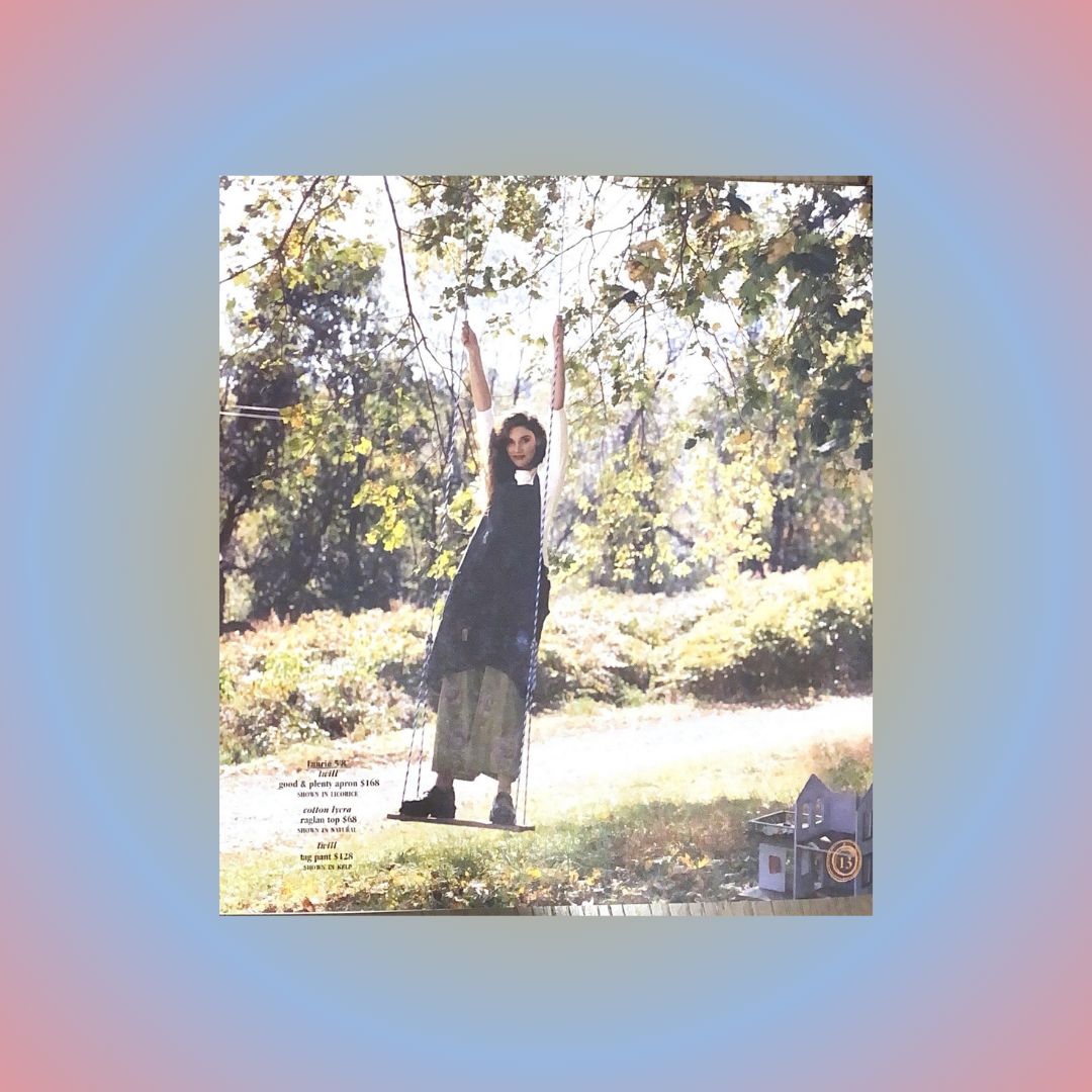 A Playful model swings on a tree swing sporting the Apron Jumper from the spring 2001 collection made from twill. Shadowing is distinctive with bright contrasts in the field of grass and tress. Her arms are up holding the ropes.