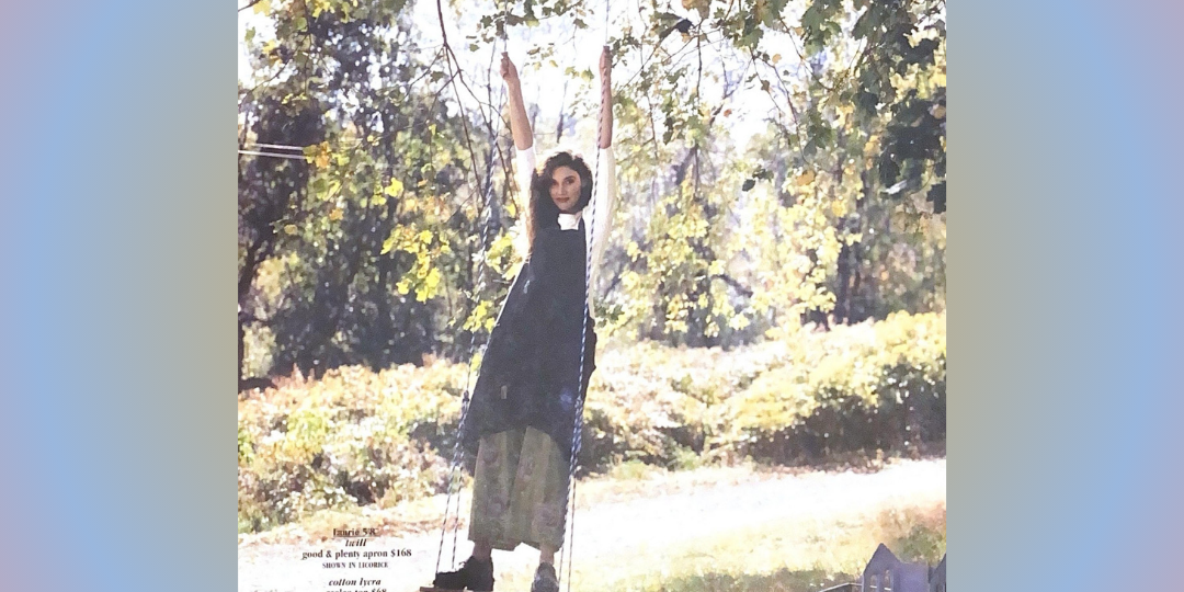 A Playful model swings on a tree swing sporting the Apron Jumper from the spring 2001 collection made from twill. Shadowing is distinctive with bright contrasts in the field of grass and tress. Her arms are up holding the ropes.