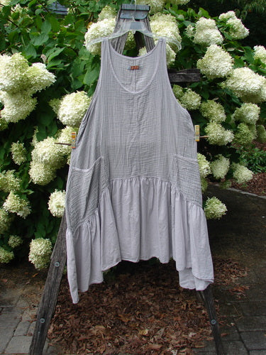 Barclay Gauze Batiste Summer Frock Jumper Stripe Light Stone Size 1, a light stone sleeveless dress with fine vertical stripes, a gathered skirt, and front patch pockets. The gauzy fabric hangs from a wooden display against dense white hydrangeas and dark greenery.