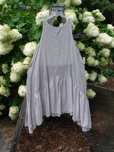 Barclay Gauze Batiste Summer Frock Jumper Stripe Light Stone Size 1 in light stone, a sleeveless gauze batiste dress with fine vertical stripes, a round neckline, and a gathered, flared hem. It hangs centered on a wooden rack amid dense white hydrangeas and green foliage.