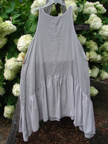 Barclay Gauze Batiste Summer Frock Jumper Stripe Light Stone Size 1 in light stone, a sleeveless gauze batiste dress with fine vertical striping, a gathered lower panel, and a loose, airy drape. It hangs against dense green foliage and white blossoms, with soft natural light emphasizing the fabric’s crinkled texture.