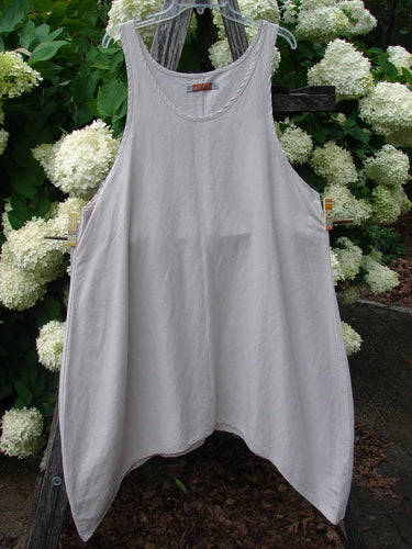 Barclay Hemp Silk Everyway Pinafore in unpainted palest peach, size 1, shown as a sleeveless, loose A-line garment with a rounded neckline and wide straps. The soft hemp-silk fabric hangs in a smooth, airy drape, with a muted peach tone against white hydrangeas and weathered wood.