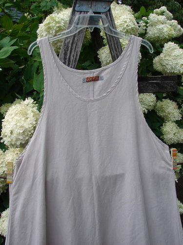 Barclay Hemp Silk Everyway Pinafore in unpainted palest peach, size 1, shown as a sleeveless slipover dress with a wide V-neck and soft drape. The lightweight fabric has a matte, airy texture, hanging from a clear hanger against green foliage and pale hydrangeas.