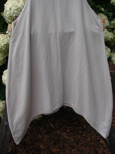 Barclay Hemp Silk Everyway Pinafore in unpainted palest peach, size 1, drapes in a loose, tent-like silhouette with a soft matte texture and gently curved hem. The lightweight fabric falls in pale peach-gray folds against an outdoor backdrop of greenery and blossoms.