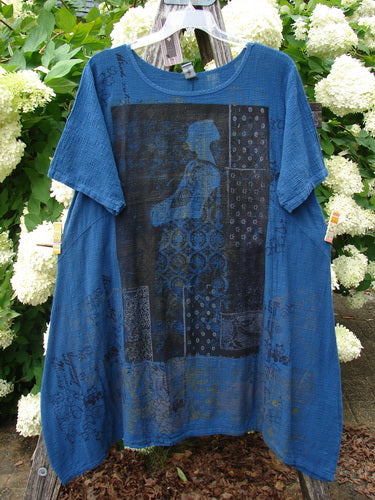 Barclay NWT Textured Gauze Chaya Tunic Window Meditation in indigo, Size 1, hangs from a clear hanger in a loose, short-sleeve silhouette. The textured gauze fabric shows a dark graphic window-meditation print across the front, with a sheer, airy drape against dense green foliage and white flowers.