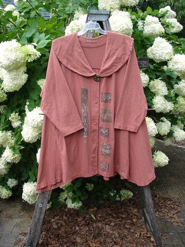 Bluefishfinder.com 1994 Wanderer's Jacket Continuous Garden Monrovian Brick Size 2 in muted brick pink, a loose, cape-like jacket with a wide rounded collar, button front, and mixed patterned panels stitched down the center. The soft fabric hangs from a hanger before white hydrangeas and dense green foliage.