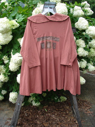 Bluefishfinder.com 1994 Wanderer's Jacket Continuous Garden Monrovian Brick size 2, a brick-pink long-sleeve jacket with a wide sailor-style collar and a loose, draped silhouette. The soft-looking fabric hangs from a wooden stand, while a small decorative chest motif adds muted detail against the dense white hydrangeas and green foliage behind it.