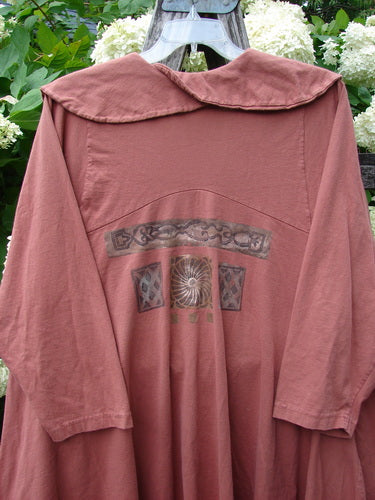 Bluefishfinder.com 1994 Wanderer's Jacket Continuous Garden Monrovian Brick Size 2 in muted brick pink, a loose long-sleeve garment with a wide rounded collar and soft drape. The back shows a faded central graphic print, hanging outdoors against leafy greenery.