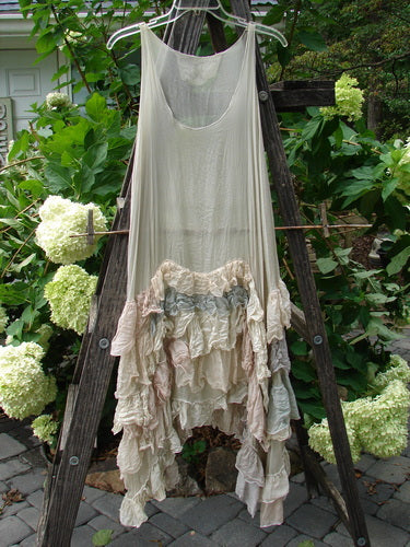 Magnolia Pearl Linen Voile Ruffle Pinafore Gray Silver Pastels OSFA displayed on a wooden ladder in an outdoor garden setting, highlighting its elegant design and delicate fabric details.