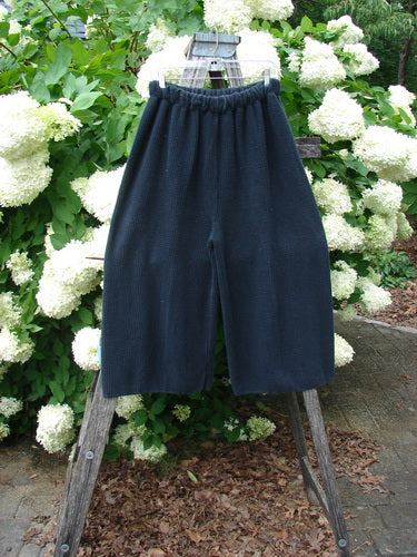 Bluefishfinder.com 1993 Thermal Waffle Wide Leg Crop Pant in unpainted black, size 1, shown with a textured waffle knit, gathered elastic waist, and cropped wide legs. The dark fabric hangs from a wooden ladder against white hydrangeas and green foliage.
