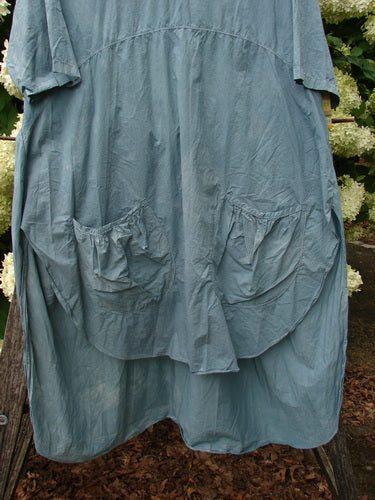 Barclay Percale Key Pocket Hi Low Dress in mottled blue cloud, size 0, with a crinkled percale texture, short sleeves, oversized front key pockets, and a softly curved hi-low hem. The dress hangs against an outdoor backdrop of greenery and wood, its muted blue-gray fabric catching gentle natural light.