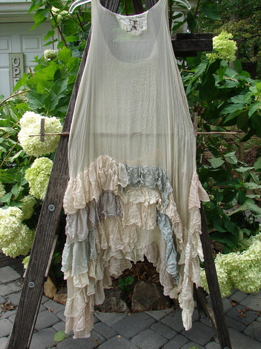 Magnolia Pearl Linen Voile Ruffle Pinafore in Gray Silver Pastels draped on a wooden ladder, showcasing its delicate ruffles and elegant design.