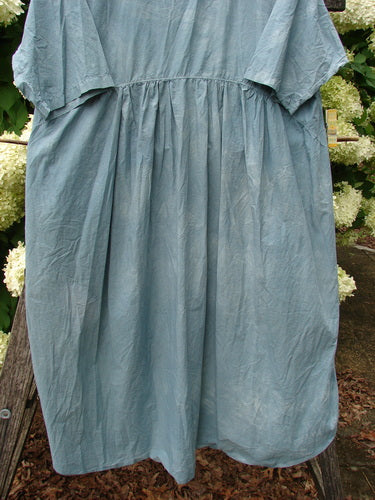 Barclay Percale Key Pocket Hi Low Dress in mottled blue cloud, size 0, with a loose gathered bodice and soft, crinkled percale texture. The hi-low hem drapes in airy panels, creating a muted blue silhouette against a floral backdrop.