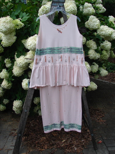 1990 The Tier Dress Polka Diamond Pink Peach OSFA in soft pink-peach fabric, a sleeveless tiered dress with a gathered ruffle hem, small dot accents, and teal patterned bands across the bodice and lower skirt. It hangs centered on a wooden stand against white hydrangeas and dark greenery, with muted natural light softening the pastel palette.