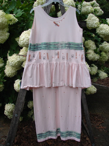 1990 The Tier Dress Polka Diamond Pink Peach OSFA in a pale pink-peach sleeveless silhouette, with a tiered ruffled waist and a soft, lightweight drape. Teal geometric bands and tiny scattered motifs add contrast, while the dress hangs against leafy white hydrangeas and weathered wood.