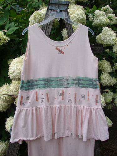 1990 The Tier Dress Polka Diamond Pink Peach OSFA, a pale pink sleeveless dress with a scoop neckline, tiered ruffled hem, and a wide mint-green geometric band across the waist. The soft fabric hangs from a clear hanger against dark wood and white hydrangeas.