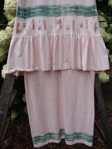 1990 The Tier Dress Polka Diamond Pink Peach OSFA, a pink-peach tiered dress with a loose, gathered ruffle at the waist and a straight lower panel. The soft fabric shows a teal patterned band near the hem and a pale geometric print across the upper tier.