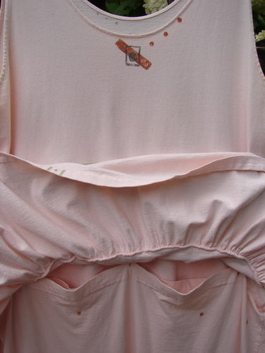 1990 The Tier Dress Polka Diamond Pink Peach OSFA in soft pink-peach fabric, shown in a close-up that reveals a sleeveless bodice, gathered tier seams, and a loose drape with small printed diamond-and-polka details. The matte textile fills the frame in layered folds, with muted blush tones and a gentle, natural light.