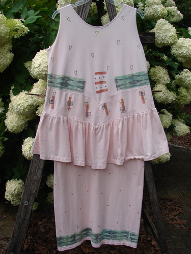 1990 The Tier Dress Polka Diamond Pink Peach OSFA in soft pink peach, a sleeveless tiered dress with a loose A-line shape, tiny diamond and polka-dot motifs, teal horizontal bands, and small printed bottle-like accents. Lightweight fabric falls over a darker wooden backdrop, with diffused outdoor light softening the pastel palette.
