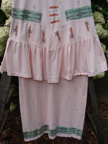 1990 The Tier Dress Polka Diamond Pink Peach OSFA in soft pink-peach fabric, shown with a tiered ruffled waist and a long straight skirt. The dress features small scattered dots and teal-and-coral geometric bands near the hem, hanging outdoors against greenery.
