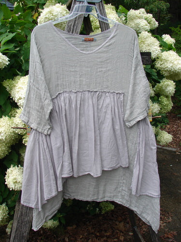 Barclay Gauze Batiste Petal Urchin Flounce Dress in unpainted rice paper, size 1, with a loose layered silhouette, soft gauze texture, and wide flounced sleeves. The pale gray-white dress hangs from a clear hanger, its airy drape set against dense green foliage and white hydrangea blooms.