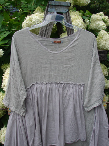 Barclay Gauze Batiste Petal Urchin Flounce Dress in unpainted rice paper, size 1, with a soft gray gauze bodice, gathered flounce waist, and loose short sleeves. The lightweight dress hangs on a clear hanger outdoors against green foliage and pale hydrangea blooms.
