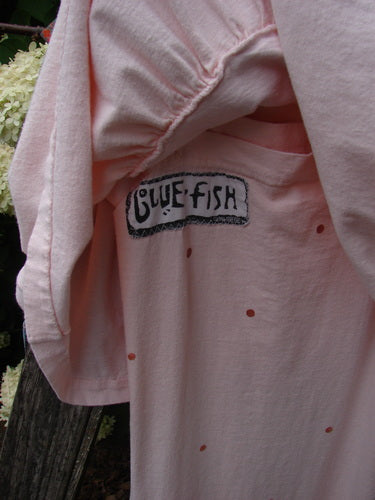 1990 The Tier Dress Polka Diamond Pink Peach OSFA in soft pink peach fabric, with tiny polka dots, gathered tiered seams, and a visible Blue Fish label at the neckline. The loose garment fills the frame in close-up, with muted natural light and a dark, textured background edge.