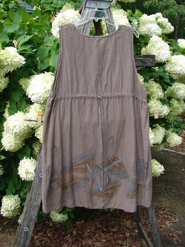 Barclay Hemp Cotton Silk Voile Venetian Pinafore Dragonfly in mocha, size 0, hangs from a wooden frame in front of white hydrangeas and green foliage. The sleeveless dress has a soft, textured voile finish, a gathered waist, and a subtle dragonfly print near the hem.