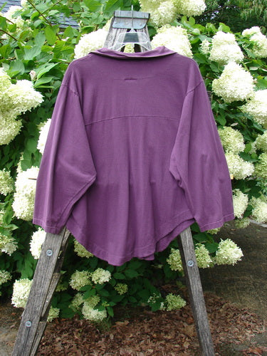 Barclay Wood Button Workshirt in unpainted plum berry, size 0, shown in a rich purple tone with a relaxed drape, wide three-quarter sleeves, and a soft crew neckline. The fabric hangs from a weathered wooden ladder, set against dense white hydrangeas and green foliage.