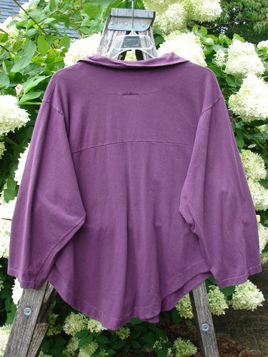 Barclay Wood Button Workshirt in unpainted plum berry, size 0, shown from the back with a soft draped collar, three-quarter sleeves, and a gently curved hem. The matte fabric hangs in a loose, layered silhouette against weathered wood and pale white blossoms.
