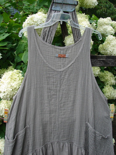 Barclay Gauze Batiste Summer Frock Jumper Stripe Sand Stone Size 1, a gray sleeveless dress with fine vertical stripes and a soft gauze-batiste texture. It hangs on a clear hanger before green foliage and white blooms, with a gathered lower section and roomy patch pockets.