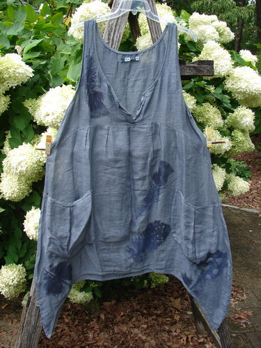Barclay Linen Tuesday's Dress in Guinea Feather Grey Storm, size 0, is a loose sleeveless linen dress with a deep V neckline and asymmetrical hem. The soft gray fabric is layered with darker indigo feather-like dye marks and hangs from a hanger against leafy white hydrangeas.