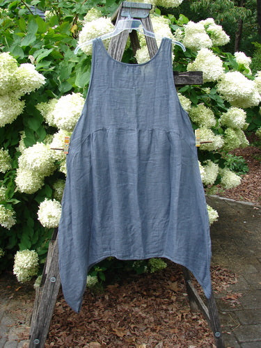Barclay Linen Tuesday's Dress Guinea Feather Grey Storm size 0, a sleeveless linen dress in muted blue-gray with a soft crinkled texture, gathered waist, and uneven hem. It hangs from a clear hanger outdoors against white hydrangeas and weathered wood.