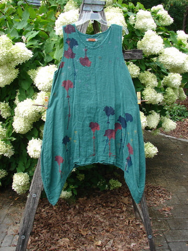Barclay Linen Round Bottom Pinafore Cloud Burst Jade in size 0, a teal-green linen pinafore with a round bottom hem and loose, sleeveless silhouette. Plum and navy floral prints trail across the front, hanging from a wooden stand against white hydrangeas.