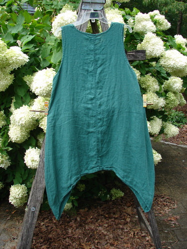 Barclay Linen Round Bottom Pinafore in Cloud Burst Jade, size 0, hangs from a wooden stand with a sleeveless A-line silhouette, rounded hem, and softly crinkled linen texture. The teal fabric is framed by white hydrangeas and greenery in natural outdoor light.